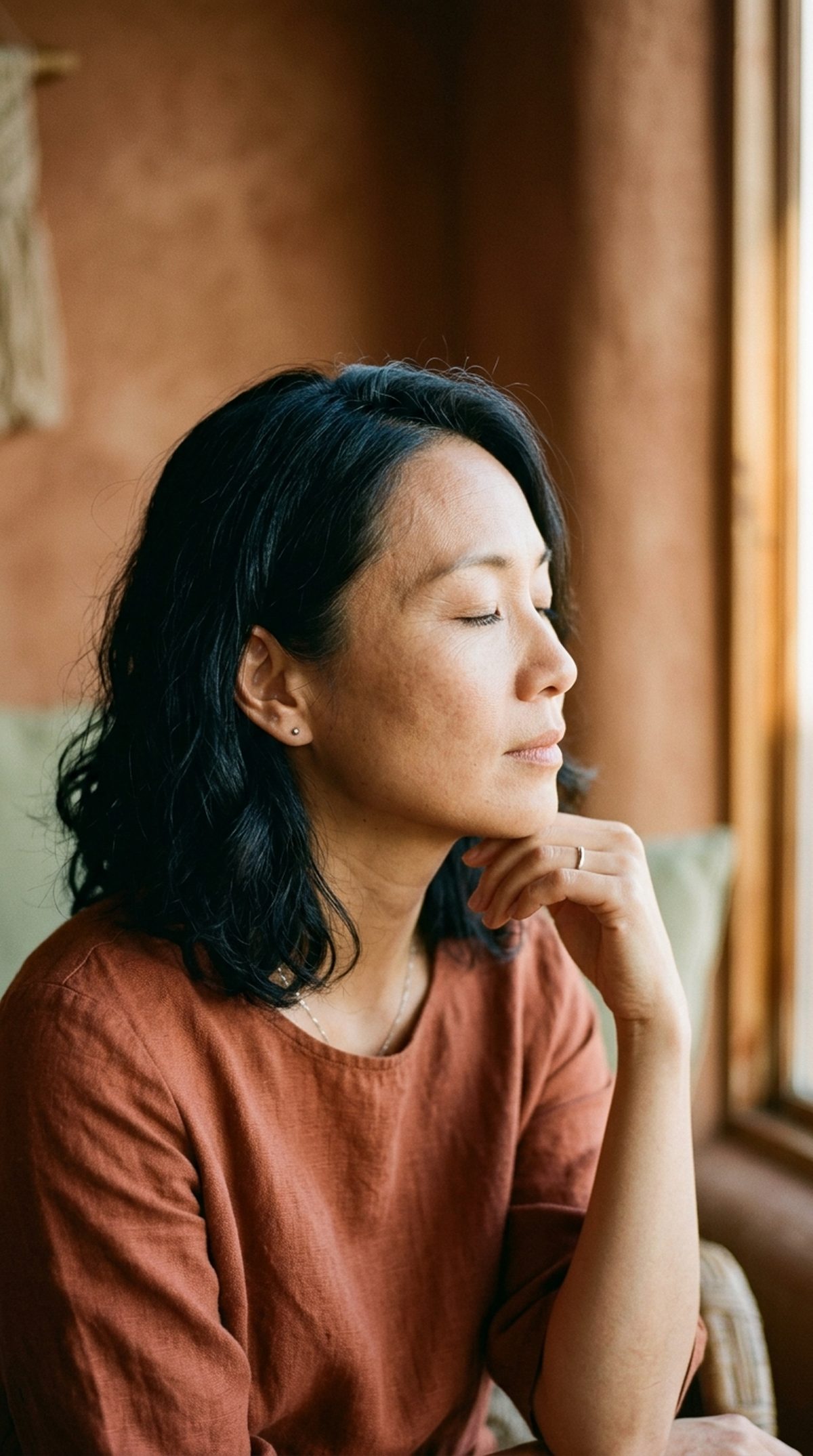 Asian woman in her 40s, eyes closed, hand near her jaw, warm afternoon light.
